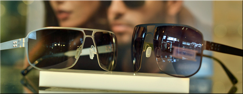 A focused close-up photograph of two pairs of high-performance polarized sunglasses, one silver-framed and one black-framed, on a white display pedestal at Optima Optometry.