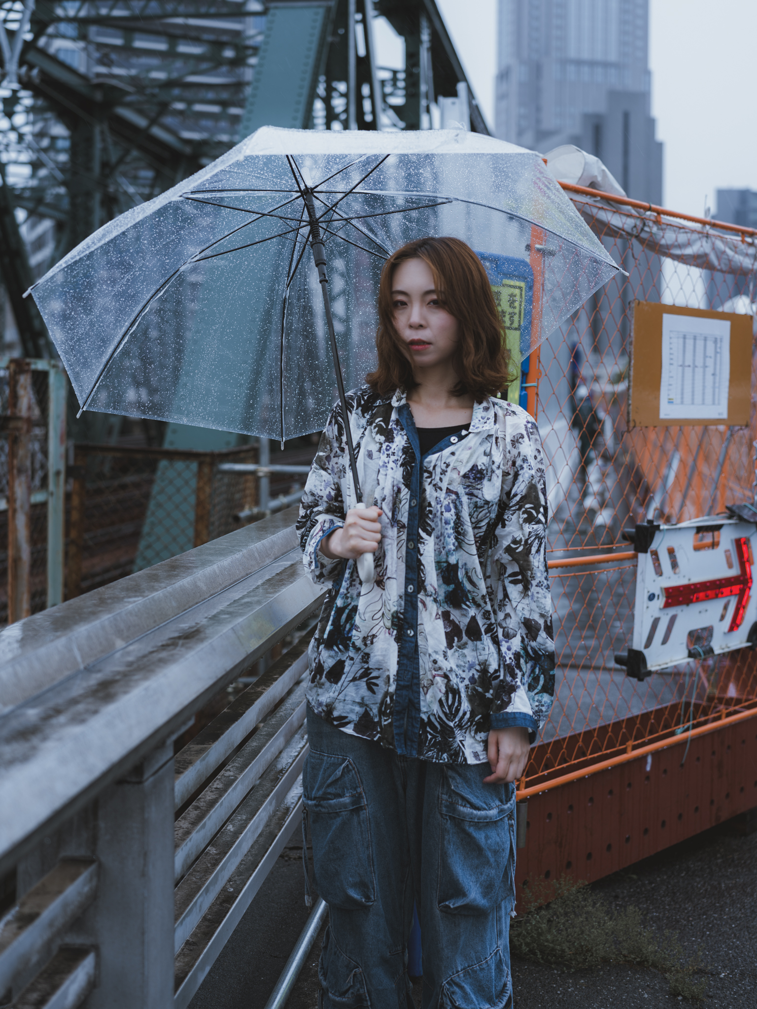 Japanese model Koishi showcasing personal street style wardrobe choices within a moody, industrial urban landscape during a rainy-day fashion editorial around Hankyu Nakatsu Station in Osaka.