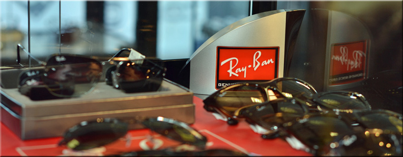 A close-up photograph of a curated display featuring a collection of Ray-Ban designer aviator and wayfarer sunglasses with a red and silver Ray-Ban brand logo stand, custom-photographed at Optima Optometry in Santa Clara.